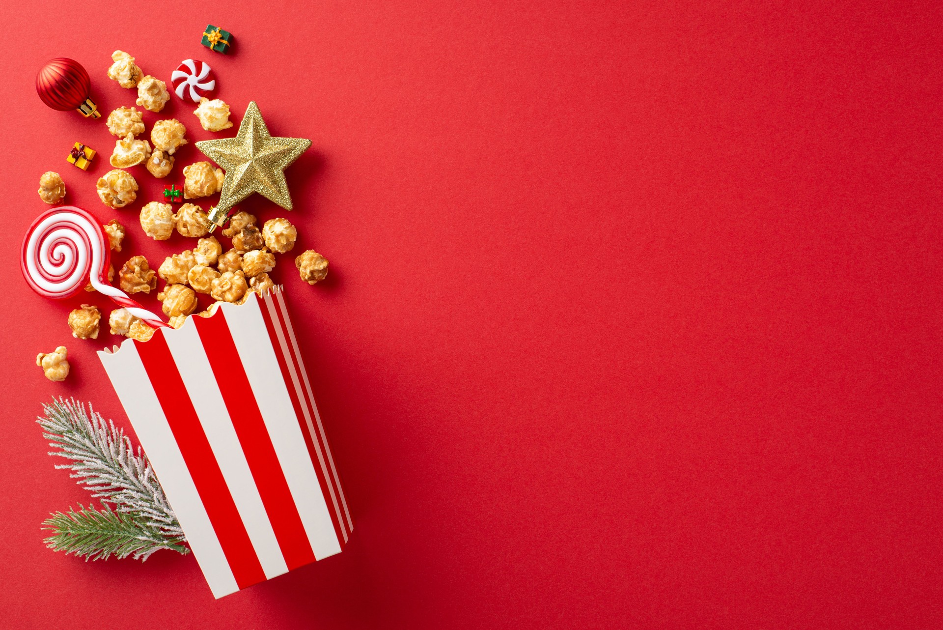 Deck the halls for New Year's film night: striped cardboard bucket with delicious popcorn, baubles, star decor, festive lollipop, candies, frosted fir branch, and a red backdrop with text space