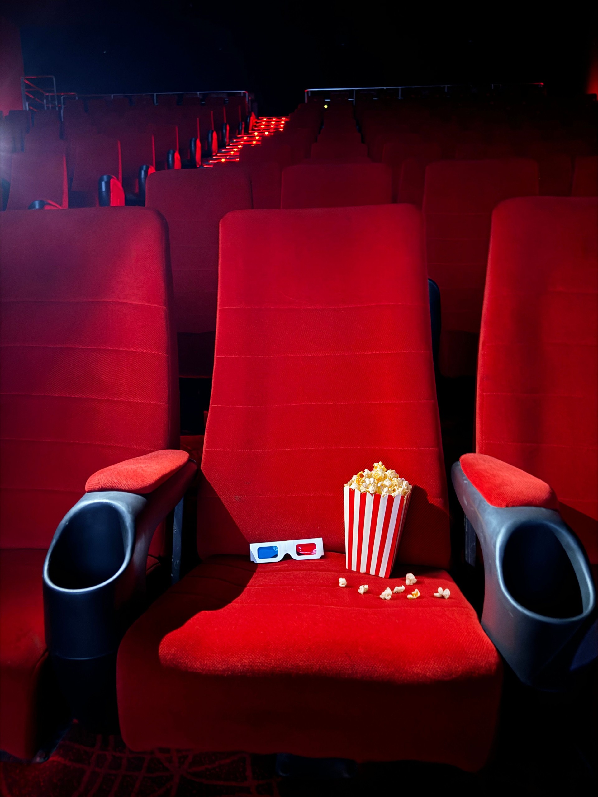 Image of striped carton of popcorn and 3D glasses in cinema theatre seat surrounded by spilled popped kernels, disposable drink cup and drinking straw in cup holder, rows of empty, red velvet cinema seats, focus on foreground
